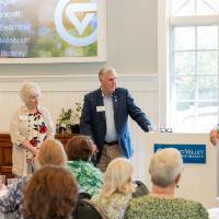 Jim Scott with microphone, addressing crowd while President and Mrs. Haas as while as President Mantella stand up by podium with him